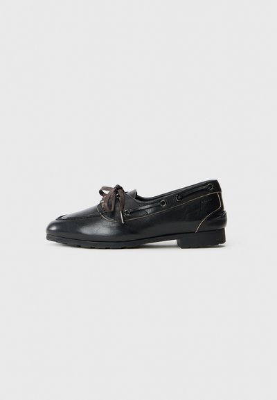 Mocassins pour hommes en cuir noir avec des lacets marron, des œillets latéraux et une semelle en caoutchouc basse sur un fond gris clair uni.