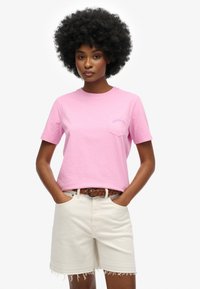 Lyserød bomulds t-shirt med korte ærmer, rund halsudskæring og en brystlomme med broderet tekst. Sammen med hvide denimshorts.