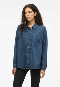 Camicia in jeans oversize di colore blu scuro, con chiusura frontale a bottoni, taschino singolo sul petto e colletto classico. Texture morbida e vestibilità rilassata.