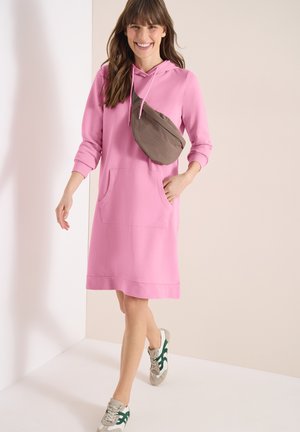Kobieta uśmiechająca się, ubrana w różową sukienkę z kapturem, szaro-zielone sneakersy oraz brązową torebkę na ramię, idąca w pomieszczeniu o neutralnych kolorach.