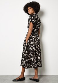 Schwarzes Kleid mit beigem Blattdruck, kurzen Ärmeln, geraffter Taille, knielanger Rock und Seitentaschen, kombiniert mit schwarzen flachen Schuhen.