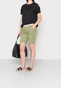 Top corto nero a maniche corte in tessuto testurizzato, pantaloni verdi, cintura bianca e borsa tote nera. Sandali con cinturini larghi e suole in legno.