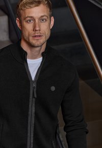 Svart zip-up stickad jacka med hög krage, prydd med en diskret logotyp på bröstet, tillsammans med en vit t-shirt under.