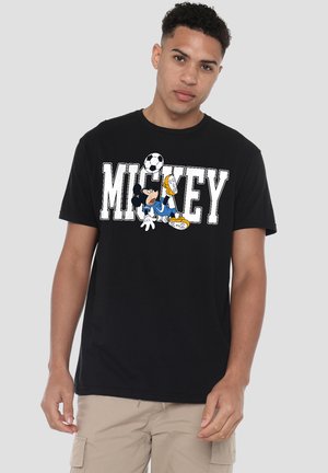 Young man wearing black T-shirt with "MICKEY" text and Mickey Mouse playing soccer graphic, paired with beige cargo shorts, neutral background.