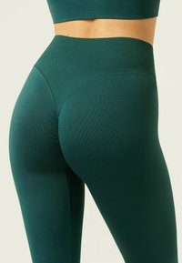 Mörkgröna högmidjade leggings gjorda av ett slätt, elastiskt material, med en konturerad söm och en sömlös midjebandsdesign.