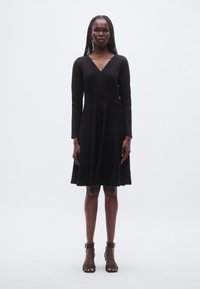 Mustast pikkade varrukatega kleit V-kaelusega, istuva ülakehaga ja lehvikukujulise seelikuga. Sile kangas peene tekstuuriga. Kandjal on mustad plokktaldadega sandaalid.