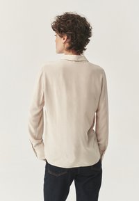 Camicia beige a maniche lunghe con una vestibilità leggermente rilassata, colletto e polsini abbottonati, abbinata a jeans scuri. Texture liscia.