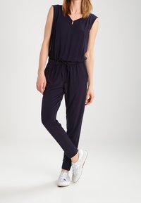 Mono azul marino con mangas cortas, escote en V y cintura ajustable con cordón; combinado con zapatillas plateadas. Tela suave, diseño de pierna ajustada.