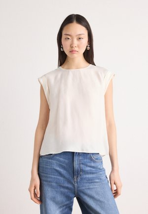 Top senza maniche di un beige chiaro, realizzato in un tessuto morbido e fluido. Collo rotondo, caduta morbida e una leggera lucentezza. Abbinato a jeans blu classici.
