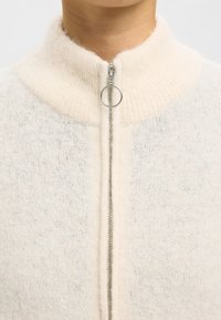 Jersey de cremallera color blanco roto con cuello alto, hecho de un material suave. Cuenta con una cremallera plateada con una pestaña circular para tirar y un acabado texturizado.