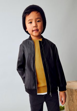 Giacca di pelle nera con chiusura a zip, polsini e fondo a coste. Indossata sopra un maglione senape. Bambino con un berretto lavorato a maglia nero, sorridente.