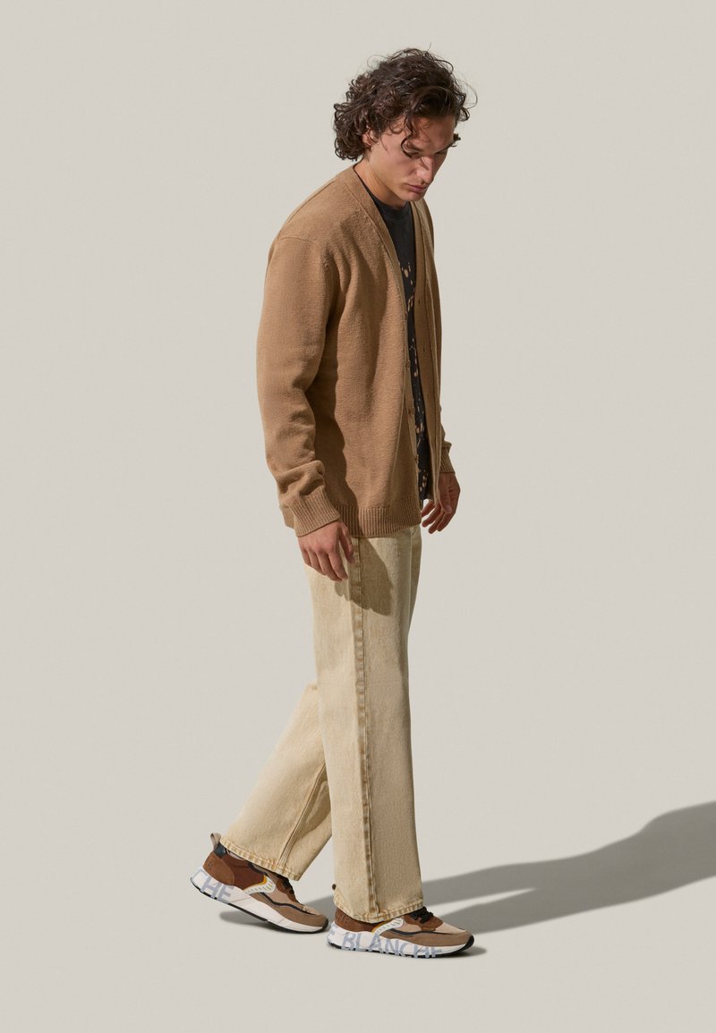 Cardigan in maglia marrone, t-shirt grafica nera, jeans a gamba larga crema e sneakers marroni/crema. I tessuti texture migliorano il look informale.