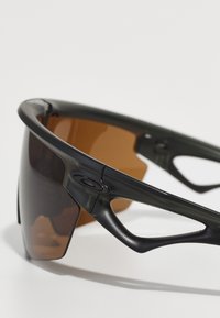 Schwarze Sonnenbrille mit sportlichem Design, featuring einen gebogenen Rahmen, braun getönte Gläser und auffällige Ausschnitte an den Bügeln.