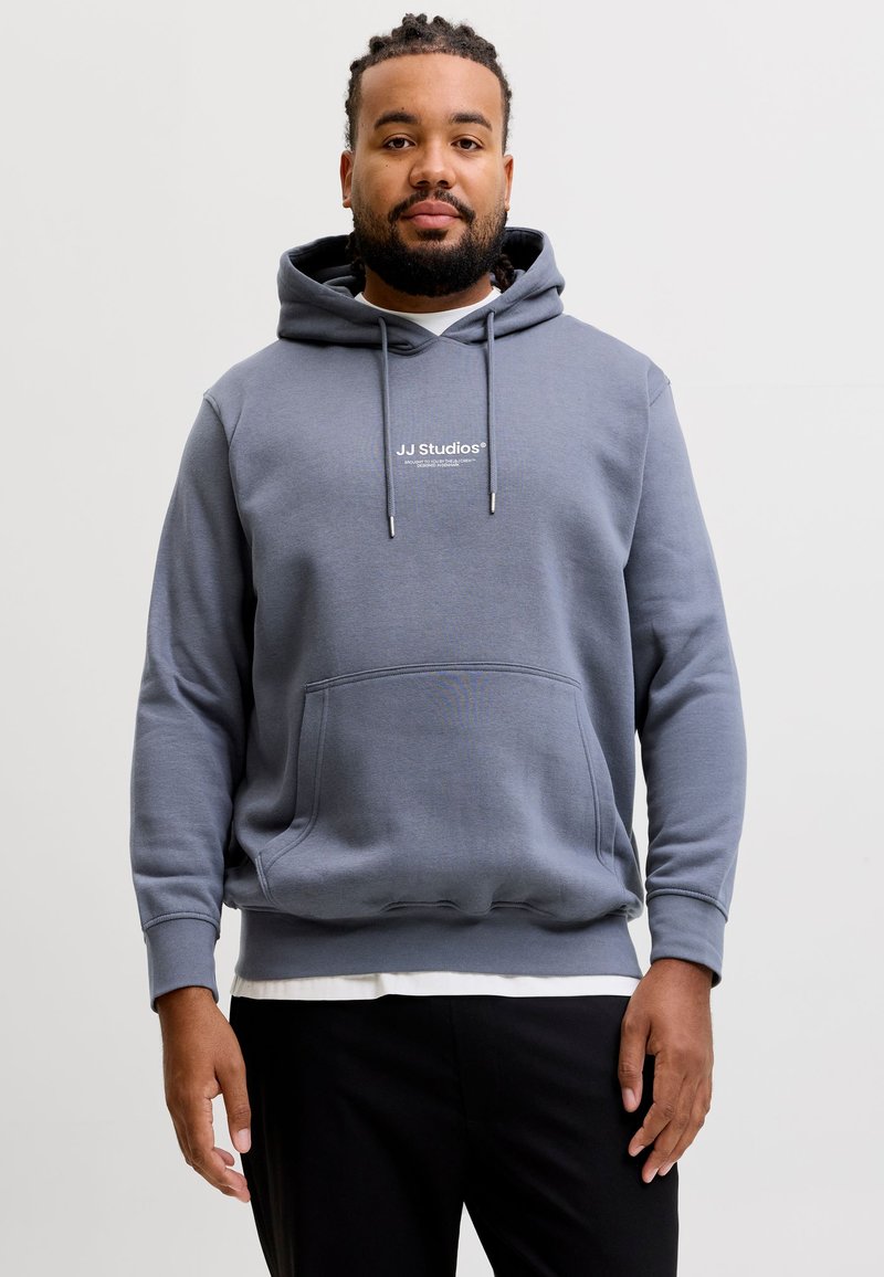 Homme avec barbe et cheveux tressés portant un sweat à capuche gris JJ Studios et un pantalon noir, debout devant un fond clair uni.