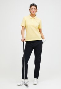 Polo shirt leggero giallo chiaro con maniche corte, colletto piatto e tre bottoni. Abbinato a pantaloni neri e scarpe da golf bianche, con in mano un bastone da golf.