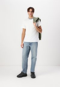 T-shirt blanc avec le logo "Lacoste" noir au col, jean bleu clair et baskets à motifs sombres. Sweat à capuche vert drapé sur un bras.