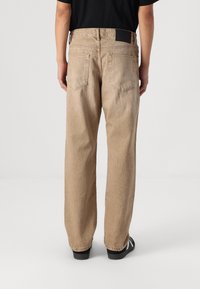 Beige wijde jeans van een textuurstof, met twee achterzakken en een zwart merklabel aan de tailleband, gecombineerd met zwarte sandalen.