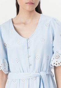 Abito azzurro chiaro con scollatura a V, maniche corte a sbuffo, ricami floreali e chiusura con bottoni. Presenta una cintura annodata in vita.