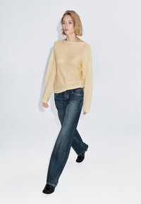 Frau, die geht und einen beigen Pullover mit langen Ärmeln, dunkelblaue Jeans mit weitem Bein und schwarze Schuhe vor einem schlichten weißen Hintergrund trägt.