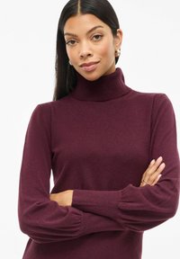 Bordeauxfarbener Rollkragenpullover mit gerippten Bündchen, figurbetontem Design und weicher Textur. Der Stoff wirkt leicht und glatt.