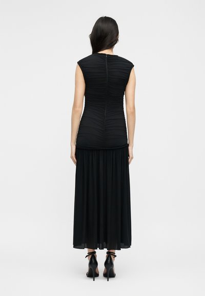 Γυναίκα με μακριά σκούρα μαλλιά που φοράει μαύρο αμάνικο τοπ με σκισίματα και μαύρη ρέουσα μακριά φούστα, όρθια με την πλάτη, με μαύρα ψηλοτάκουνα σανδάλια.