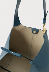 Μπλε τσάντα χειρός από δερμάτινη υφή με εσωτερικό σε μπεζ χρώμα, ανοιχτό πάνω μέρος, χρυσό μαγνητικό κούμπωμα και ευρύ λουράκι ώμου με χρυσές λεπτομέρειες.