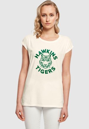 Kremfarget t-skjorte med korte ermer som har et grønt grafisk design av et tigeransikt med teksten "HAWKINS TIGERS" over og under.