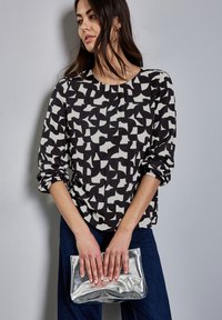Top a maniche lunghe con motivo astratto bianco e nero, scollo a crew e polsini arrotolati. Il modello tiene una borsa a tracolla argentata lucida.