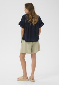 Blusa navy con maniche a volant e bordo in pizzo, abbinata a pantaloni corti verde chiaro. Modella che indossa slip-on intrecciati beige, rivolta di spalle rispetto alla macchina fotografica.