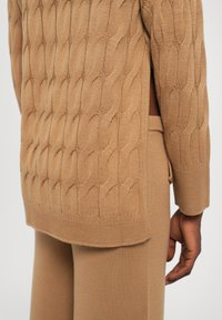 Pullover in maglia cable-knit color sabbia con un motivo testurizzato e ondulato e spacchi laterali. Le maniche lunghe e l'orlo arrotondato completano il design.