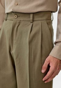 Olijfgroene op maat gemaakte broek in een gestructureerde stof, met voorplooien, riemlussen en een knoopsluiting bij de taille.