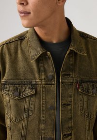 Τζιν μπουφάν σε ελιά πράσινο με κουμπιά μπροστά, δύο τσέπες στο στήθος, αντιθετική ραφή και κλασικό γιακά. Φορεμένο πάνω από μαύρο πουκάμισο.