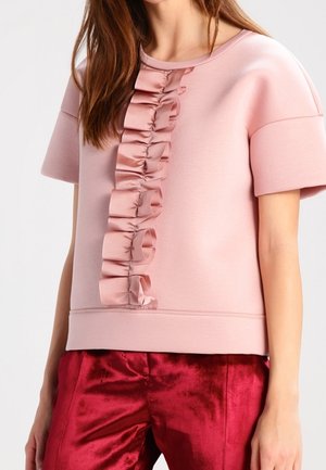 Frau trägt ein hellrosa kurzärmeliges Oberteil mit vertikalen, gerafften Satinbesätzen und glänzende rote Samthose.