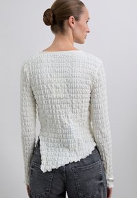 Kobieta z włosami upiętymi w kok, ubrana w teksturowany biały top z długim rękawem i ciemnoszare dżinsy, zwrócona plecami na jasnoszarym tle.