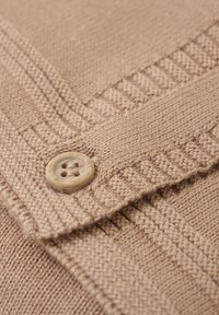 Beige stickad tyg med ett strukturerat mönster och en rund, krämfärgad knapp med fyra hål. Stygn är detaljerade och konsekventa.