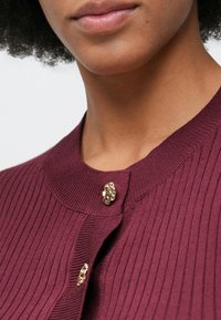 Close-up de uma pessoa a usar um cardigã ribado em maroon com botões texturizados em dourado únicos abotoados no pescoço.