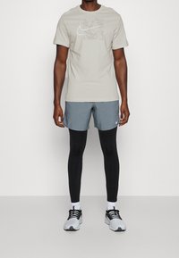 Jasnoszara koszulka Nike z grafiką logo, zestawiona z niebieskimi szortami, czarnymi leginsami i czarno-białymi butami sportowymi. Prosty, sportowy design.