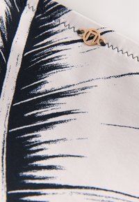 Tissu blanc avec imprimé feuille de palmier noire, couture en zigzag noire et petit bijou métallique circulaire doré fixé près du bord.