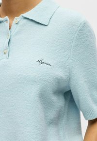 Polo à manches courtes bleu clair en tissu doux et texturé avec col et patte de boutonnage à trois boutons. Présente un accent brodé "élégance".