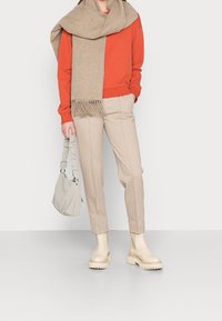 Beige Chinos, oranger Sweatshirt und ein beiger Schal mit Fransen. Cremige Stiefeletten und eine hellgraue Tasche mit gerafften Griffen.
