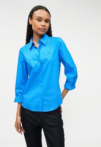 Sinine kraega nööpidega särk, mille varrukad on üles keeratud ja mis on valmistatud sileda kangaga. Rinnal on väike logo. Sobib tumedate teksadega.