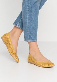 Mocasin amarillo perforado con punta redonda y suela flexible, que presenta intrincados patrones recortados y un acabado liso y texturizado.