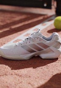 Witte tennisschoen met beige accenten op een gravelbaan dichtbij een witte grenslijn en een gele tennisbal op de achtergrond.