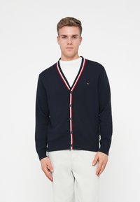 Ναυτικό ζακέτα με λευκή και κόκκινη ρίγα στο V-λαιμό, κλείσιμο με κουμπιά, πλεκτές μανσέτες και ένα μικρό λογότυπο στο αριστερό στήθος. Κατασκευασμένη από μαλακό πλεκτό υλικό.