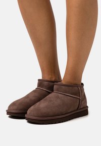 Botas de tobillo de ante marrón con puntera redondeada, detalles cosidos, una textura suave y suela de goma, mostradas en un par de piernas.