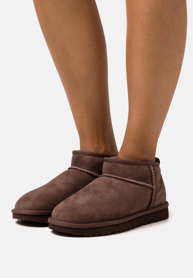 Botas de tobillo de ante marrón con puntera redondeada, detalles cosidos, una textura suave y suela de goma, mostradas en un par de piernas.