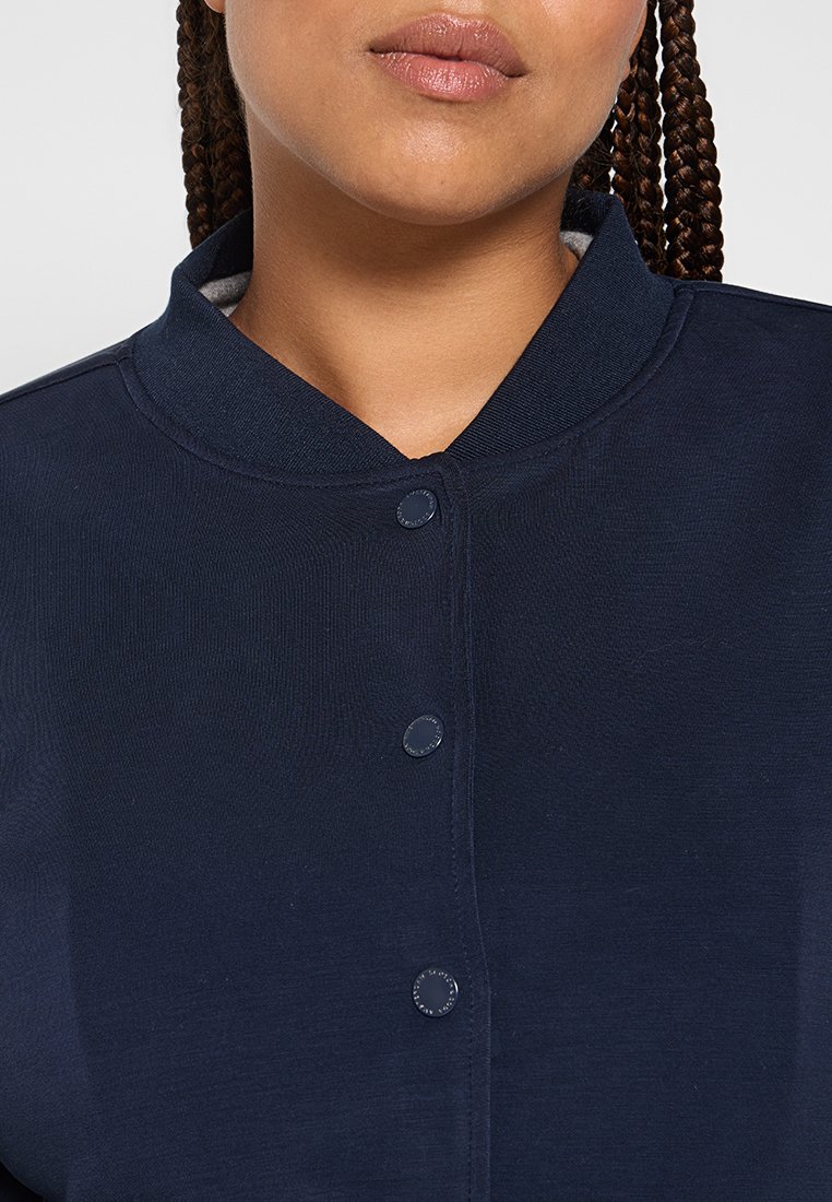 Femme aux cheveux tressés portant une veste boutonnée bleu marine avec un col côtelé, les lèvres légèrement entrouvertes, sur un fond neutre.