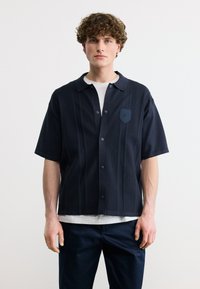 Νέος άνδρας με σγουρά μαλλιά φοράει μπλε σκούρο κοντομάνικο πουκάμισο με κουμπιά πάνω από λευκό T-shirt, σε λευκό απλό φόντο.