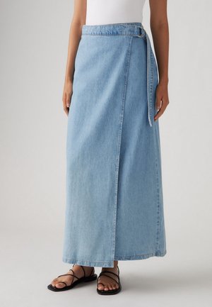 Femme portant une jupe portefeuille en denim bleu clair avec une ceinture à boucle et des sandales plates noires à brides sur un fond uni.