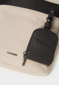 Portafoglio in pelle nera attaccato a una borsa di tessuto beige. La borsa ha una texture liscia e presenta una tracolla nera con cuciture visibili.